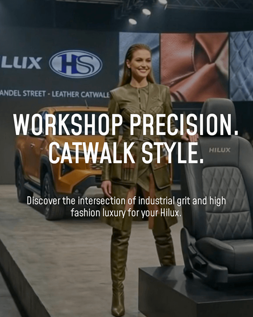 A fashion model in tailored olive green leather attire standing on a runway next to a custom dark grey quilted leather Hilux car seat with a vehicle in the background.